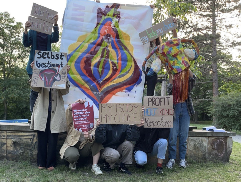 Das Foto zeigt eine Gruppe von Leute, die hinter verschiedenen Plakaten und Postern stehen. Zentral ist dabei eine große gezeichnete Vulva, um das Vulva-Plakat herum werden Plakate in die Höhe gehalten mit den Wortlauten: "Safer Sex - Safer Use", "Selbstbestimmung!", "Faires PJ!", "My Body My Choice" und "Profit heilt keine Menschen".